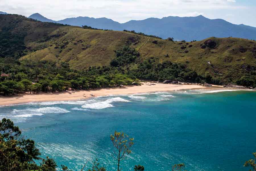 IMAGEM: a fotografia mostra uma praia de ilhabela com o mar azul, uma faixa de areia e árvores na orla. ao fundo, há um morro coberto por vegetação. FIM DA IMAGEM.