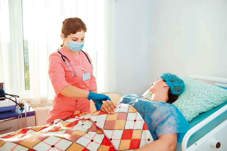 IMAGEM: uma mulher usando jaleco rosa e luvas está segurando o punho de outra mulher, que está deitada na cama de um hospital. FIM DA IMAGEM.
