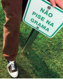 IMAGEM: Pessoa pisando em um jardim apesar da placa que mostra o desenho de um sapato atravessado por um risco vermelho indicando proibição. FIM DA IMAGEM.