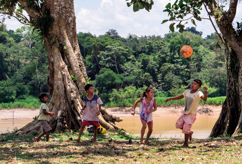 IMAGEM: a fotografia que inicia a unidade 1 mostra crianças peruanas jogando futebol ao lado de duas grandes árvores na margem de um rio. as crianças estão descalças e vestem roupas leves como bermudas e camisetas. ao fundo, do outro lado do rio, há uma floresta. FIM DA IMAGEM.