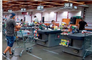 IMAGEM: caixas em um supermercado. alguns clientes aguardam na fila com seus carrinhos, outros clientes são atendidos pelos operadores de caixas. FIM DA IMAGEM.