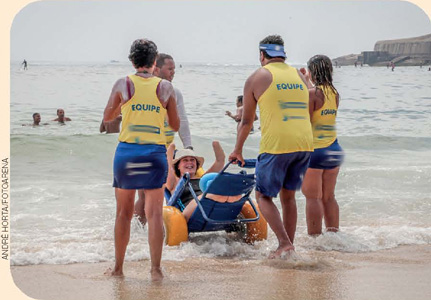 IMAGEM: pessoas usando regatas e shorts ajudam uma mulher usuária de cadeira de rodas a entrar no mar em uma cadeira adaptada. a mulher está rindo e levantando os braços. FIM DA IMAGEM.