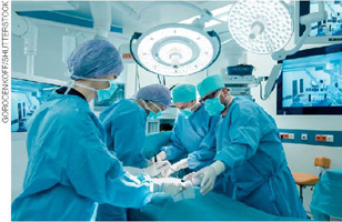 IMAGEM: médicos e enfermeiros fazendo uma cirurgia em uma sala equipada com luzes e monitores. eles usam roupas de proteção com toucas, máscaras e luvas. FIM DA IMAGEM.