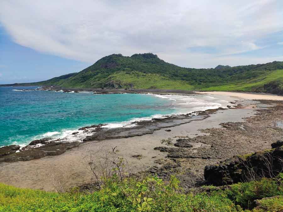 IMAGEM: a fotografia de fernando de noronha mostra o mar azul, a faixa de areia da praia, rochas na beira do mar e, ao fundo, um morro coberto por vegetação. FIM DA IMAGEM.