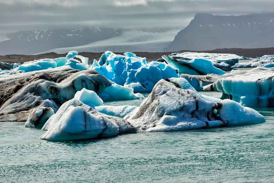 IMAGEM: grandes blocos de gelo dentro de um lago. os blocos apresentam coloração azul e preta. ao fundo, há várias montanhas entre nuvens. FIM DA IMAGEM.