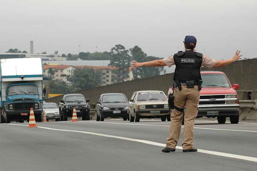 IMAGEM: uma mulher de costas usando boné e colete à prova de balas acena para alguns carros em uma estrada. FIM DA IMAGEM.