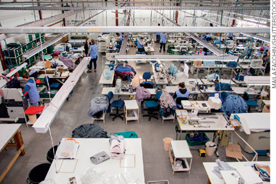 IMAGEM: vista do alto do interior de uma fábrica de roupa, com maquinário e várias bancadas. FIM DA IMAGEM.