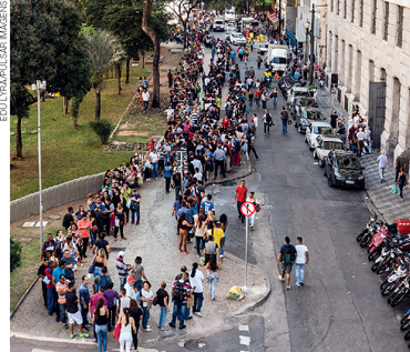IMAGEM: vista aérea de uma rua em que chama a atenção uma fila muito grande de pessoas em uma calçada. FIM DA IMAGEM.