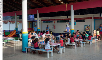 IMAGEM: pátio de uma escola na hora lanche. as crianças estão sentadas em mesas e bancos adequados ao seu tamanho. o ambiente é ordenado e limpo. FIM DA IMAGEM.