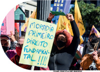 IMAGEM: fotografia de uma manifestação de rua com pessoas carregando bandeiras e cartazes. um deles diz: moradia primeiro direito fundamental!!!!. FIM DA IMAGEM.