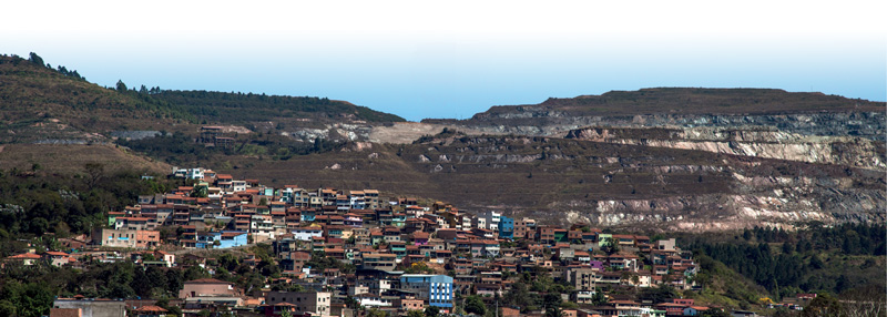IMAGEM: vista de uma cidade em uma região de planalto, com muitas casas e edifícios nas encostas. ao fundo, as montanhas estão desmatadas e recortadas pela exploração de minério. FIM DA IMAGEM.
