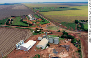 IMAGEM: vista aérea de uma região rural com extensa área de plantação cortada por estradas de terra, algumas construções com galpões e silos, e poucas casas. FIM DA IMAGEM.