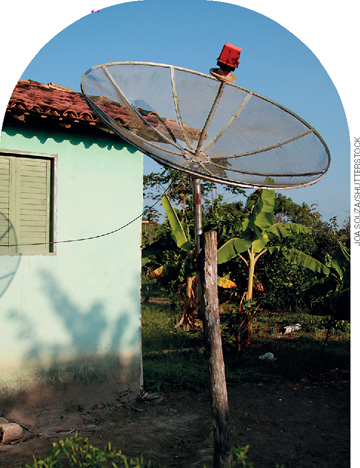 IMAGEM: uma antena parabólica ao lado de uma casa simples, na área rural. FIM DA IMAGEM.