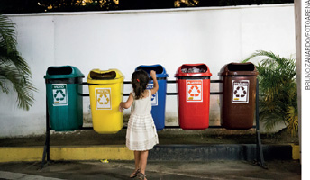 IMAGEM: uma menina pequena joga lixo em uma de cinco lixeiras para materiais recicláveis. FIM DA IMAGEM.
