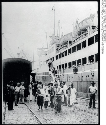 IMAGEM: fotografia antiga de um grupo de imigrantes, em que homens mulheres e crianças, com roupas de época, desembarcam de um navio. FIM DA IMAGEM.