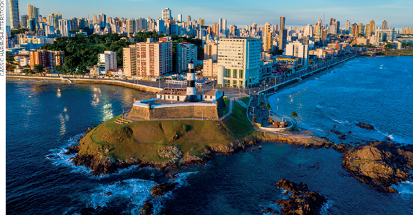 IMAGEM: vista aérea da cidade litorânea de salvador. toda a orla da praia é tomada por edifícios altos, avenidas asfaltadas, iluminadas e arborizadas. há um grande farol sobre uma fortificação em um cabo de rochas que avança pelo mar. FIM DA IMAGEM.