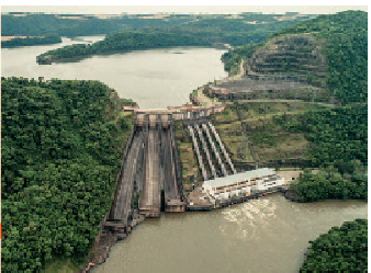 IMAGEM: Imagem aérea de uma usina hidrelétrica, que é uma estrutura de concreto com paredes quase verticais com comportas que permitem que a água represada passe pelo desnível, gerando energia. FIM DA IMAGEM.