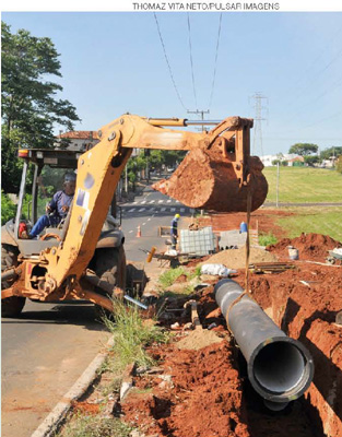 IMAGEM: uma escavadeira abre uma vala na terra ao lado de uma estrada, onde um tubo está sendo instalado. FIM DA IMAGEM.