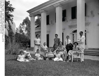IMAGEM: fotografia em preto e branco de onze pessoas, homens, mulheres e crianças, usando roupas características da década de 1950, em um jardim em frente a uma mansão. FIM DA IMAGEM.