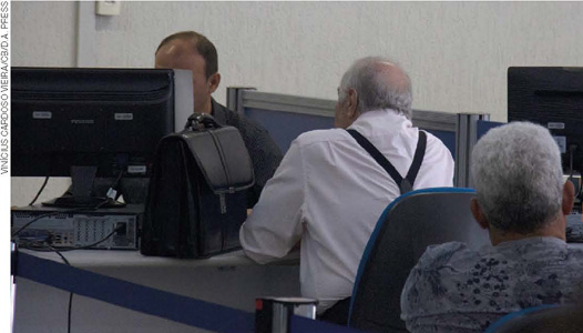 IMAGEM: um idoso está em frente a um homem adulto. ambos estão sentados em uma mesa de escritório, o adulto está atrás de um computador. FIM DA IMAGEM.