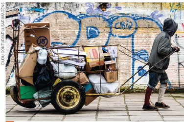 IMAGEM: em um cenário urbano, com muro pichado atrás, um catador puxa uma carroça com materiais recicláveis, como papelões. FIM DA IMAGEM.