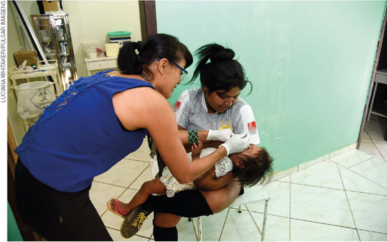 IMAGEM: uma enfermeira vacina uma criança, que está no colo da mãe. FIM DA IMAGEM.