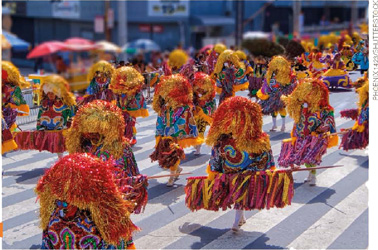 IMAGEM: participantes de um bloco de maracatu desfilam na rua. eles usam grandes adornos arredondados na cabeça, vestem roupas estampadas e levam bastões decorados com franjas coloridas. FIM DA IMAGEM.