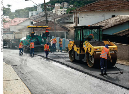 IMAGEM: um grupo de trabalhadores com roupas de proteção trabalha próximo a caminhões de pavimentação. eles alisam o asfalto recém colocado em uma rua. FIM DA IMAGEM.