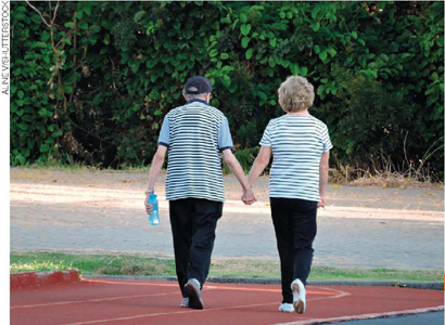 IMAGEM: um homem e uma mulher idosos caminham de mãos dadas em uma pista em área verde. FIM DA IMAGEM.