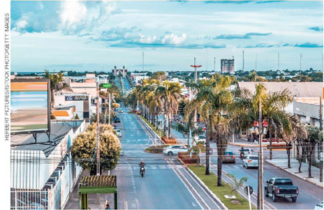IMAGEM: paisagem de uma rua com duas vias, palmeiras, carros e motos circulando, prédios comerciais e vegetação ao fundo. FIM DA IMAGEM.
