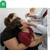 IMAGEM: 2. uma mulher segura uma criança enquanto uma enfermeira aplica vacina de gota em sua boca. FIM DA IMAGEM.