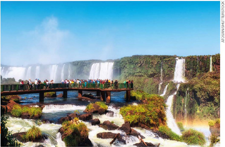 IMAGEM: a paisagem das cataratas do iguaçu com diversas cachoeiras e uma plataforma repleta de turistas. há rochas e um arco-íris. FIM DA IMAGEM.