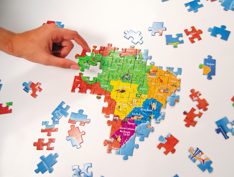 IMAGEM: uma mão monta um quebra-cabeças. há peças soltas pela mesa e a parte já montada mostra um mapa do brasil com as divisões dos estados e regiões. FIM DA IMAGEM.