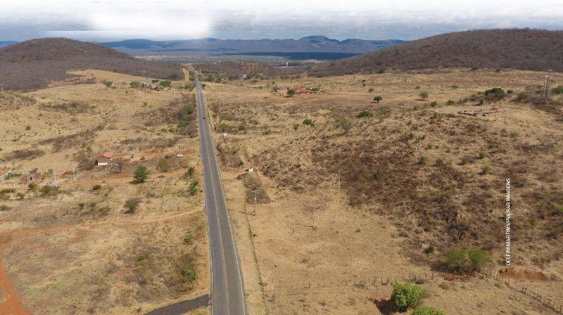 IMAGEM: vista aérea de estrada em região seca, com alguns arbustos, chão de terra e montanhas no horizonte. FIM DA IMAGEM.