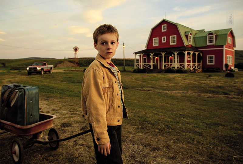 IMAGEM: abrindo ao capítulo sete, está a fotografia de um menino pequeno carregando um carrinho de reboque com rodas, com sua mala sobre ele. o menino tem um olhar triste e olha para longe. veste uma jaqueta sobre uma camisa listrada e calças compridas. ele está em frente a uma casa grande com dois andares e sótão, em um lugar remoto, descampado, com pequenas elevações ao fundo. um carro está estacionado próximo à casa. FIM DA IMAGEM.