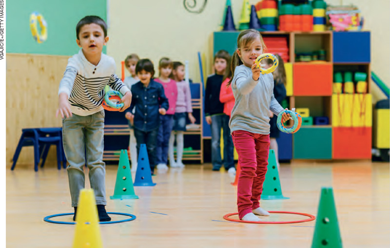 IMAGEM: crianças formam duas filas, lado a lado. elas seguram argolas em suas mãos, tentando acertar cones que estão no chão. FIM DA IMAGEM.