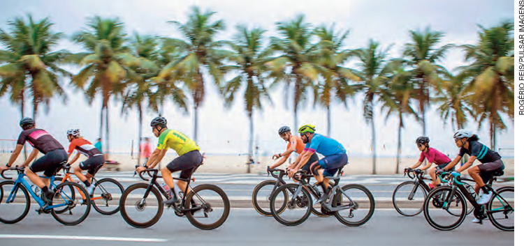 IMAGEM: um grupo de ciclistas pedala em uma avenida próxima à praia. FIM DA IMAGEM.