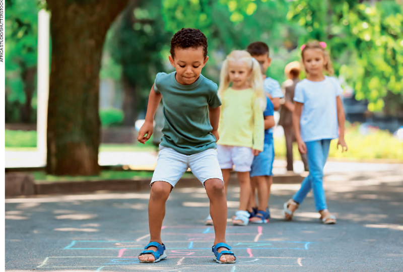 IMAGEM: crianças em fila brincam de amarelinha em um parque. FIM DA IMAGEM.