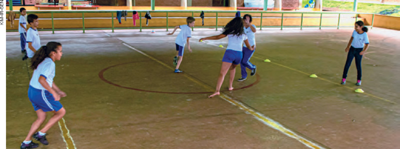 IMAGEM: crianças brincam em uma quadra esportiva, divididas em dois times, um de cada lado da quadra. no centro, um grupo se aproxima. FIM DA IMAGEM.