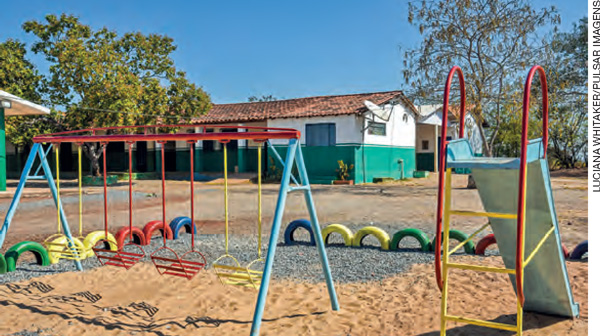 IMAGEM: parquinho de uma escola, com balanços, escorregador e pneus coloridos. ao fundo, a construção da escola e árvores. FIM DA IMAGEM.