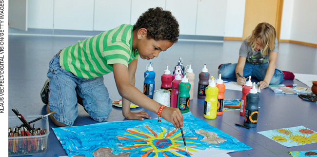 IMAGEM: duas crianças sentadas no chão fazem pinturas em grandes folhas de papel. elas usam tubos de tinta colorida e pincéis. FIM DA IMAGEM.