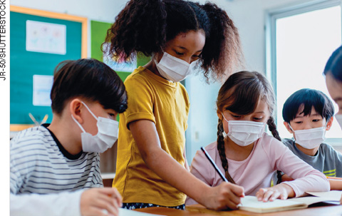 IMAGEM: cinco crianças em uma sala de aula. uma delas está de pé e escreve em um caderno enquanto as outras observam. todas usam máscaras cirúrgicas. FIM DA IMAGEM.