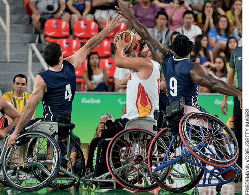 IMAGEM: atletas paralímpicos jogam basquetebol. todos eles estão em cadeiras de rodas, e disputam a bola. ao fundo, a torcida assiste ao jogo. FIM DA IMAGEM.