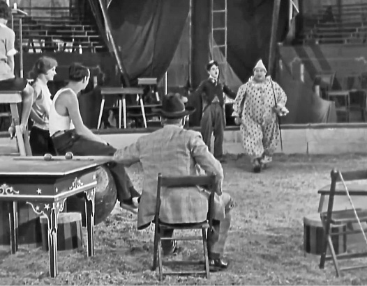 Imagem: Fotografia em preto e branco. Borda com formato de filme. No centro, Chaplin e um palhaço estão andando em um picadeiro. Na frente deles, três pessoas estão sentadas e os observando. Ao fundo, uma arquibancada vazia. Fim da imagem.