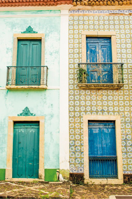 Imagem: Fotografia. Duas construções geminadas de dois andares. À esquerda, parede verde-claro com janelas verde-escuro. À direita, parede com bolinhas amarelas e janelas azul-escuro. Fim da imagem.