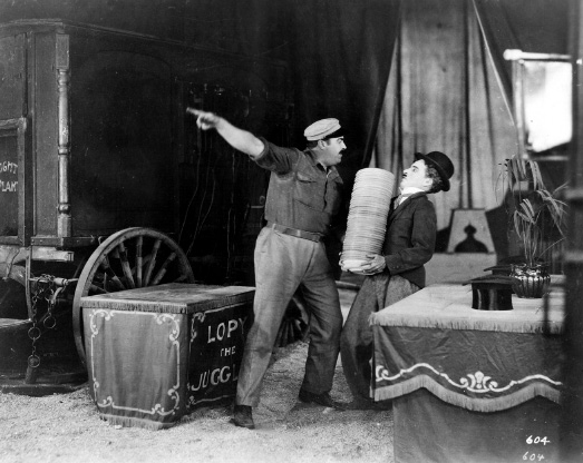 Imagem: Fotografia em preto e branco. Borda com formato de filme. No centro, Chaplin está segurando uma pilha de pratos com o corpo inclinado para trás. Ao seu lado, um homem está com o corpo inclinado para frente e o dedo indicador direito apontado para o lado. Em volta deles há objetos, uma carruagem e uma caixa.  Fim da imagem.