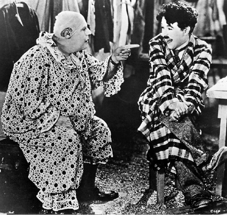Imagem: Fotografia em preto e branco. Borda com formato de filme. No centro, Chaplin com roupa xadrez está sentado e sorrindo. Ao seu lado, o palhaço está sentado e apontando para ele com o dedo indicador.  Fim da imagem.