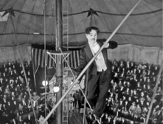 Imagem: Fotografia em preto e branco. Borda com formato de filme. No centro, Chaplin com terno está andando em uma corda suspensa e segurando um bastão comprido. Abaixo, pessoas observam sentadas em uma arquibancada.  Fim da imagem.