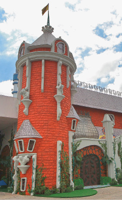 Imagem: Fotografia. Construção com formato de castelo. Na lateral há uma torre vermelha com detalhes cinza e no topo, uma bandeira. Fim da imagem.