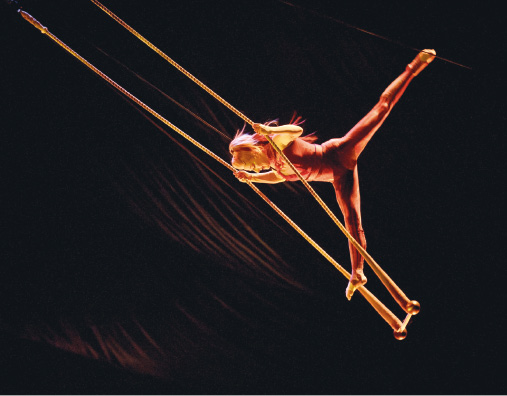 Imagem: Fotografia. Uma bailarina está pendurada em duas cordas e com a perna esquerda esticada para trás.  Fim da imagem.